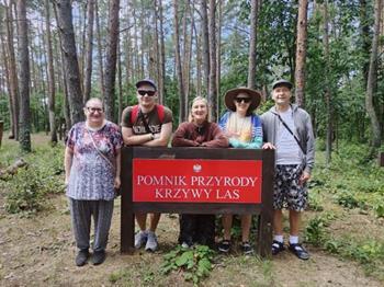 Uczestnicy opierający się o tablicę z napisem „Pomnik Przyrody Krzywy Las” na tle drzew